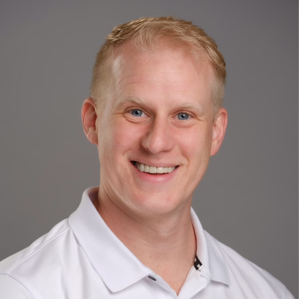 Head-and-shoulders portrait of a smiling man with short blond hair wearing a white polo shirt against a gray studio background.